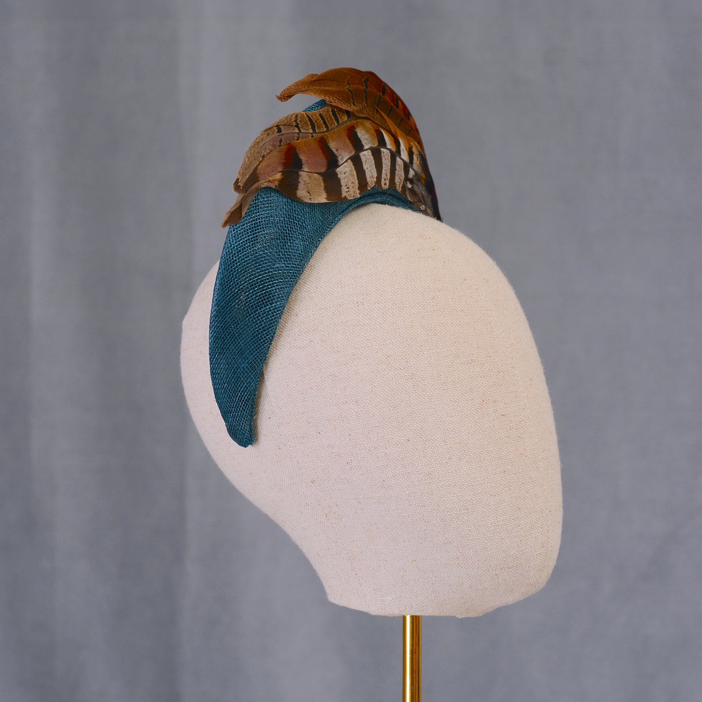 A teal headband adorned with brown and tan feathers. The feathers are arranged in layers, creating a textured appearance. The headband is displayed on a white mannequin head.