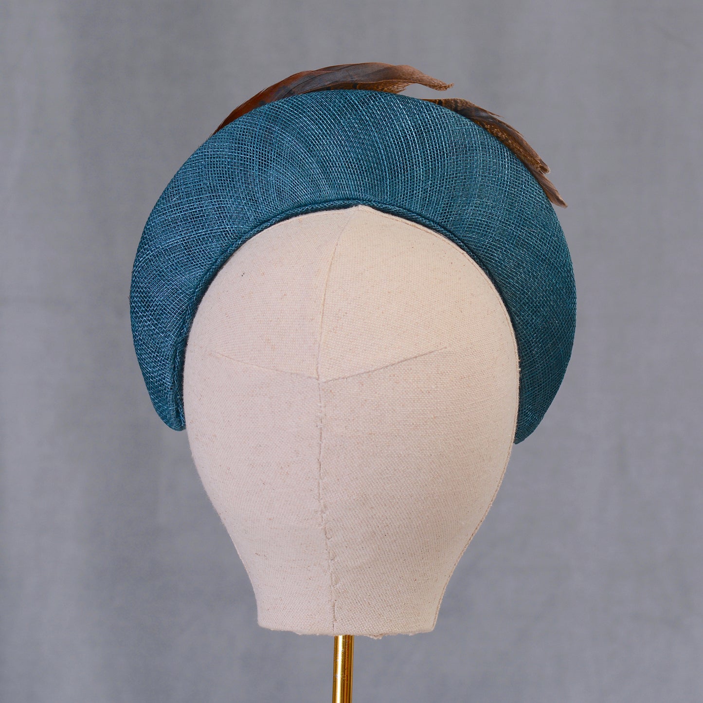 A teal hat with a curved design, made from textured straw and adorned with brown feathers. The hat is displayed on a mannequin head, highlighting its shape and style. This accessory is suitable for special occasions.