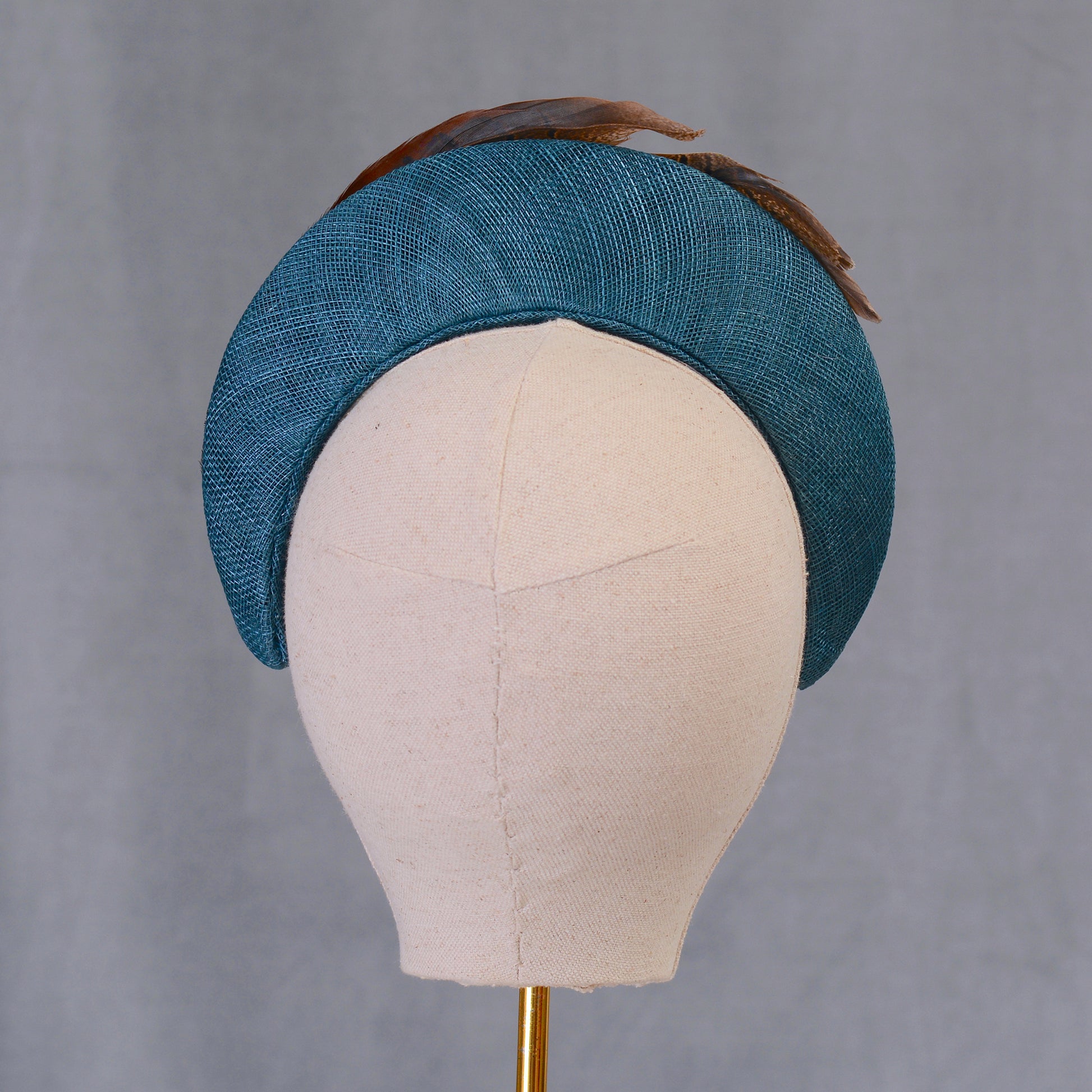 A teal hat with a curved design, made from textured straw and adorned with brown feathers. The hat is displayed on a mannequin head, highlighting its shape and style. This accessory is suitable for special occasions.
