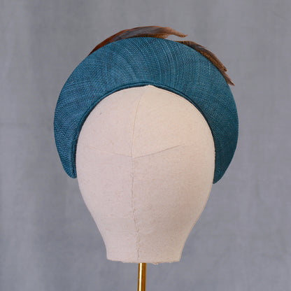 A teal hat with a curved design, made from textured straw and adorned with brown feathers. The hat is displayed on a mannequin head, highlighting its shape and style. This accessory is suitable for special occasions.