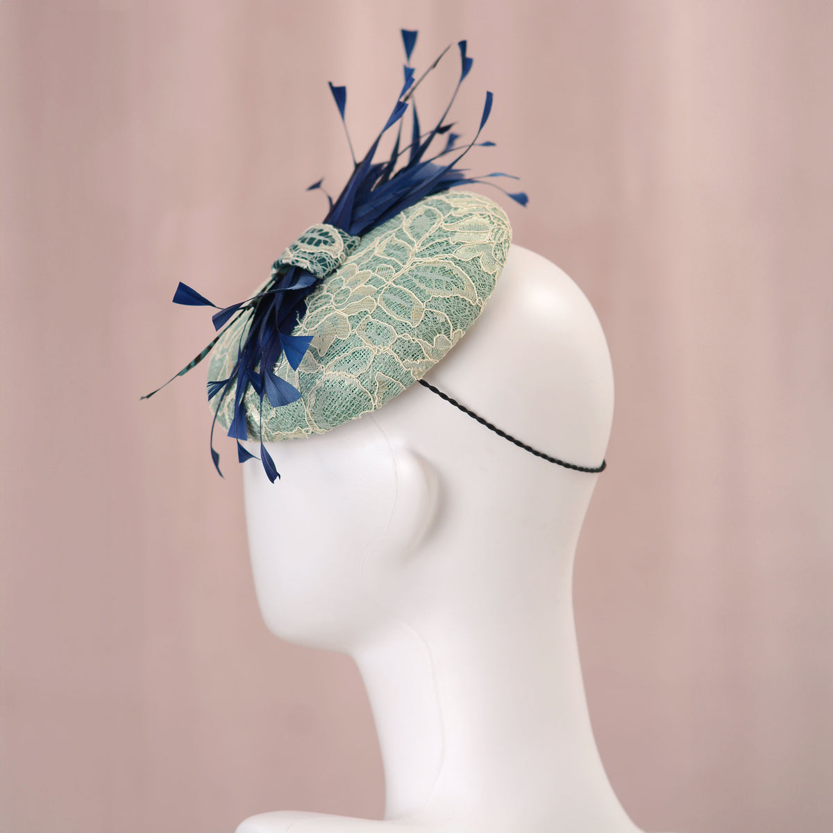 A wedding fascinator hat displayed on a white mannequin head against a soft pink backdrop. The hat features a pale turquoise green lace-embroidered circular  button base with dark blue  and teal feathers.