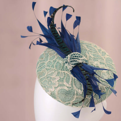 A lace fascinator hat displayed on a white mannequin head against a soft pink backdrop. The hat features a pale turquoise green lace-embroidered circular  button base with dark blue  and teal feathers.