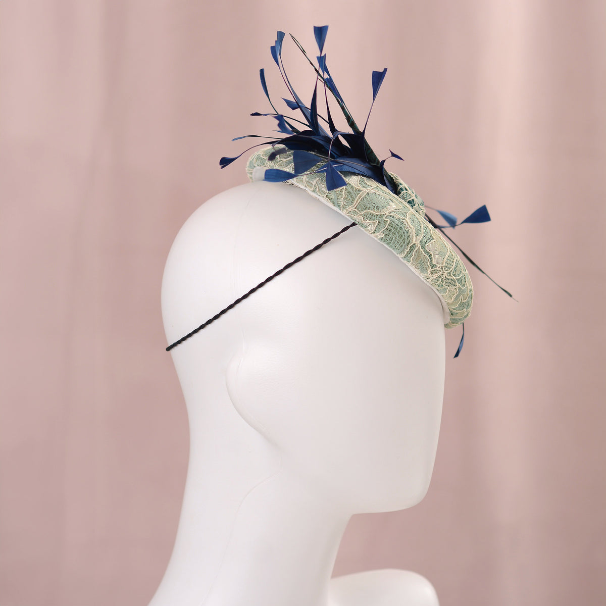 A wedding fascinator hat displayed on a white mannequin head against a soft pink backdrop. The hat features a pale turquoise green lace-embroidered circular  button base with dark blue  and teal feathers.