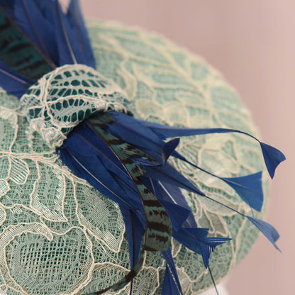 A close-up of a decorative fascinator hat featuring a pale mint green lace crown with long royal blue feathers and a patterned blue feather accent.