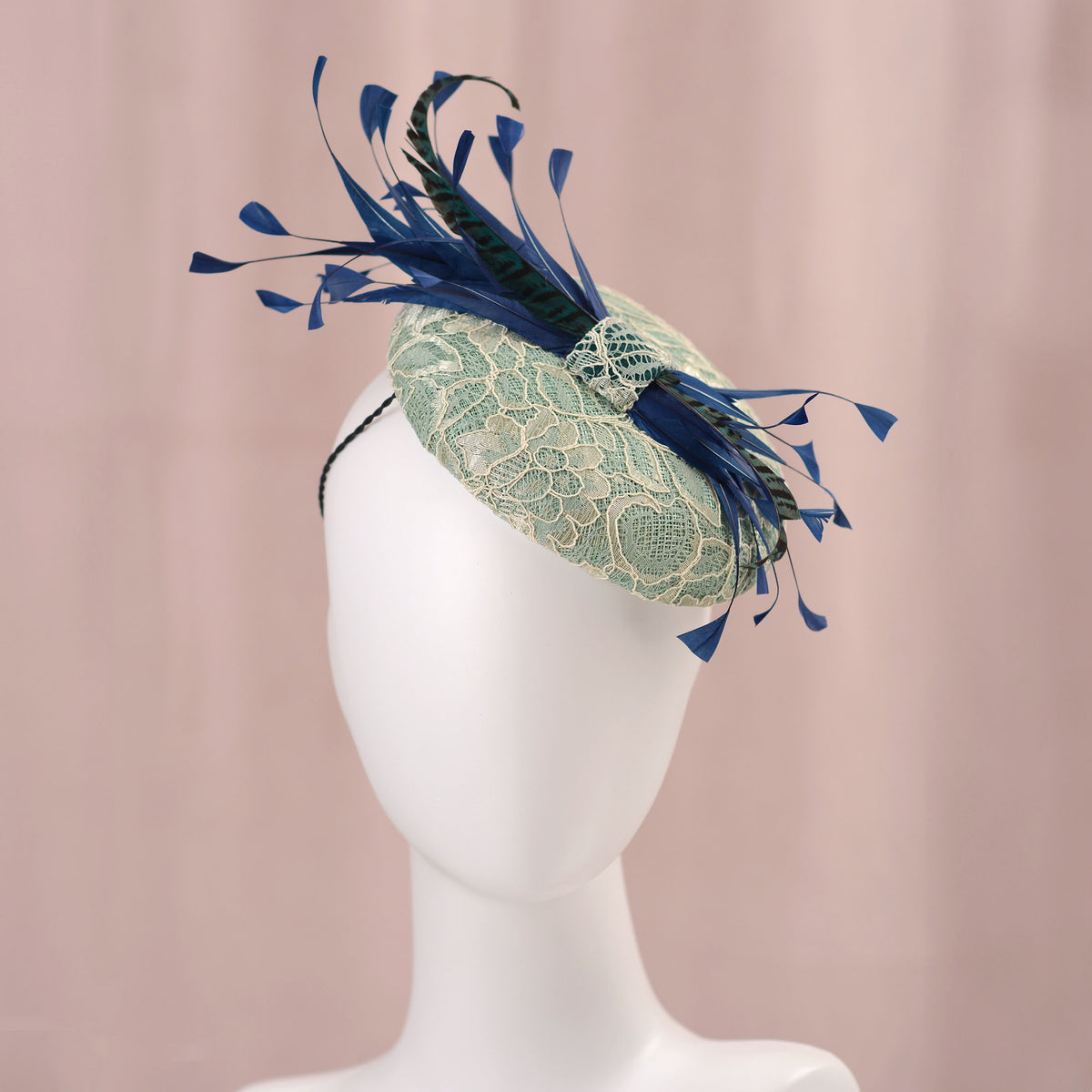 A decorative fascinator hat displayed on a white mannequin head against a soft pink backdrop. The hat features a pale turquoise green lace-embroidered circular  button base with dark blue  and teal feathers.