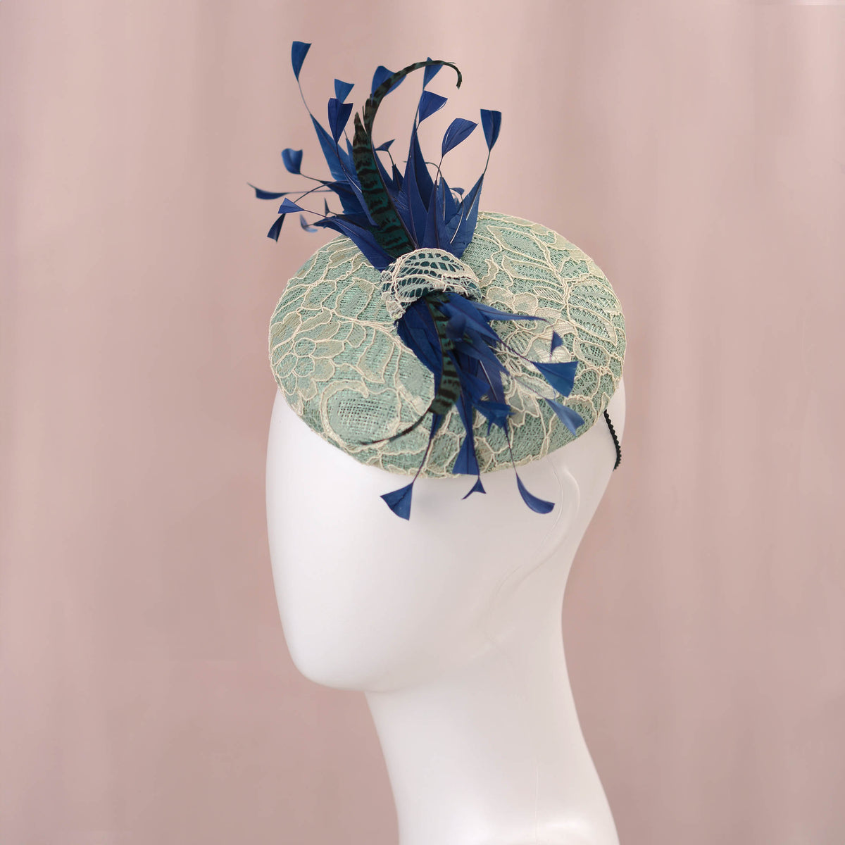 A horse racing fascinator hat displayed on a white mannequin head against a soft pink backdrop. The hat features a pale turquoise green lace-embroidered circular  button base with dark blue  and teal feathers. ideal for Royal Ascot 