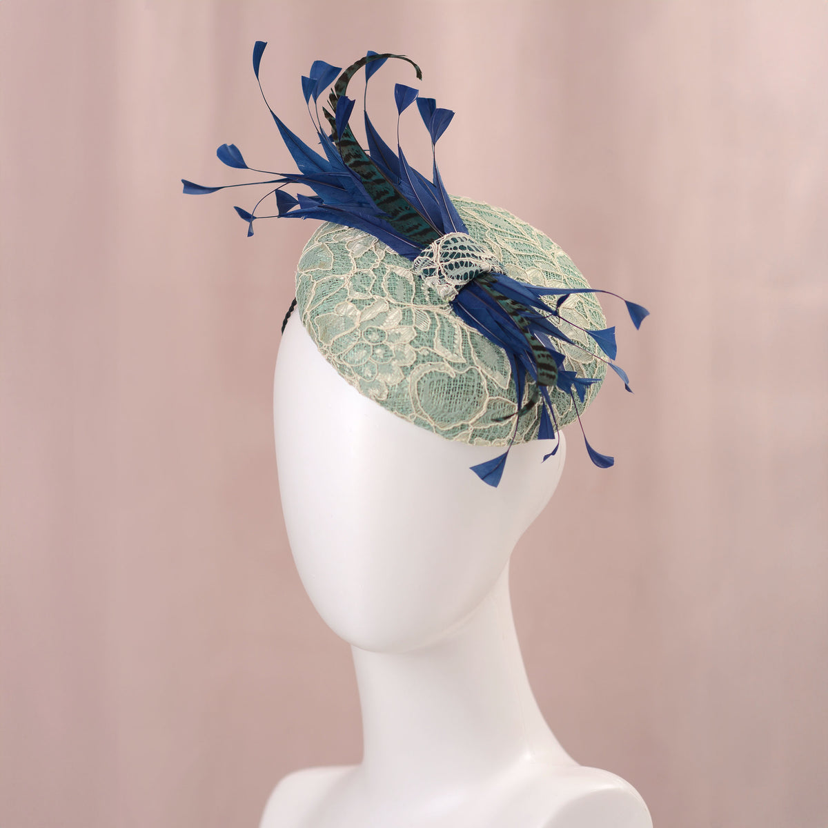 A wedding fascinator hat displayed on a white mannequin head against a soft pink backdrop. The hat features a pale turquoise green lace-embroidered circular  button base with dark blue  and teal feathers.