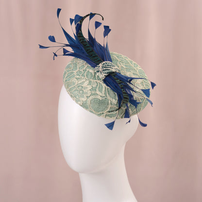 A light mint green lace fascinator hat with navy blue and teal feathers, displayed on a white mannequin head against a soft pink background.
