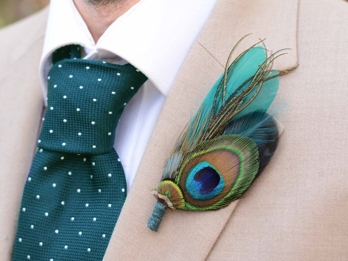 A close-up of a beige suit jacket with a white dress shirt and a dark green polka-dot tie, featuring a decorative peacock feather boutonniere pinned to the lapel.