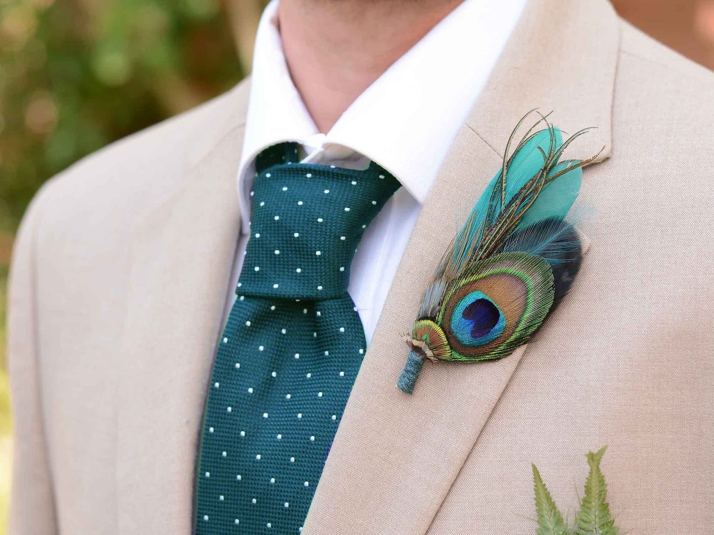 A close-up of a man's chest wearing a beige suit jacket, white dress shirt, and a dark teal polka-dot tie, with a decorative peacock feather boutonniere pinned to the left lapel.