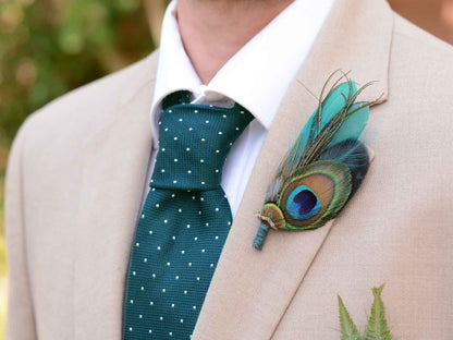 A close-up of a man's chest wearing a beige suit jacket, white dress shirt, and a dark teal polka-dot tie, with a decorative peacock feather boutonniere pinned to the left lapel.