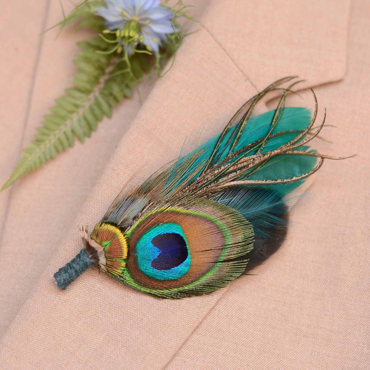 A decorative peacock feather boutonniere pin accent, pinned to a beige suit jacket with a blue flower and green fern in background.