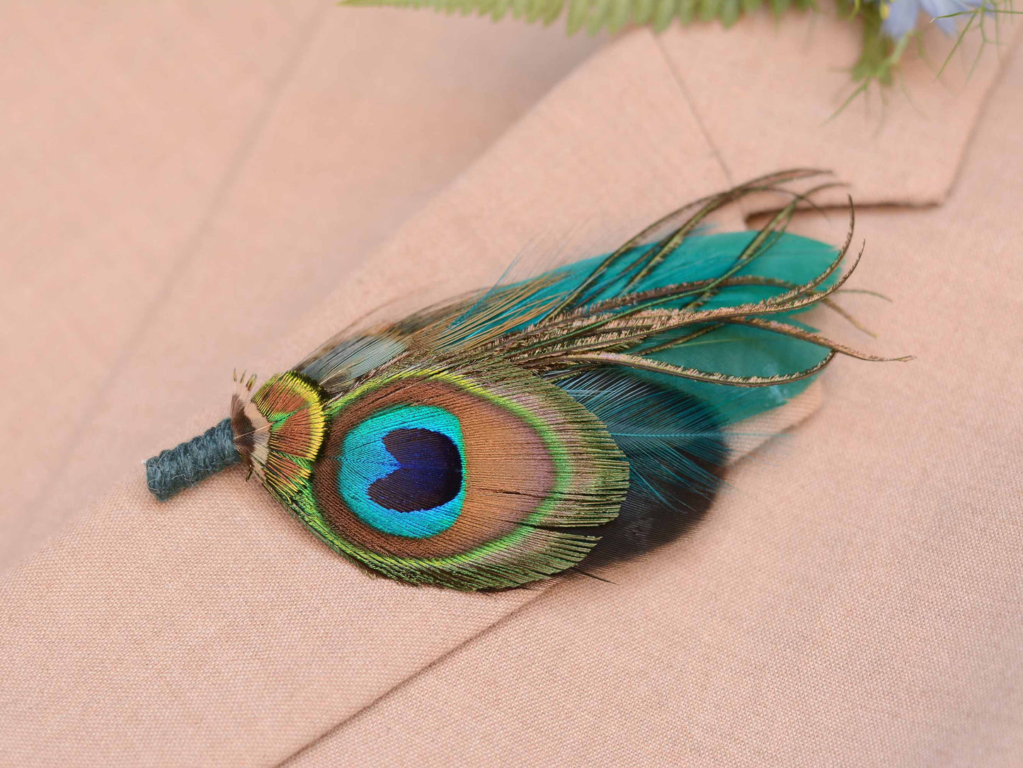 Close up of a  decorative peacock feather boutonniere pin accent, pinned to a beige suit jacket with a blue flower and green fern in background.