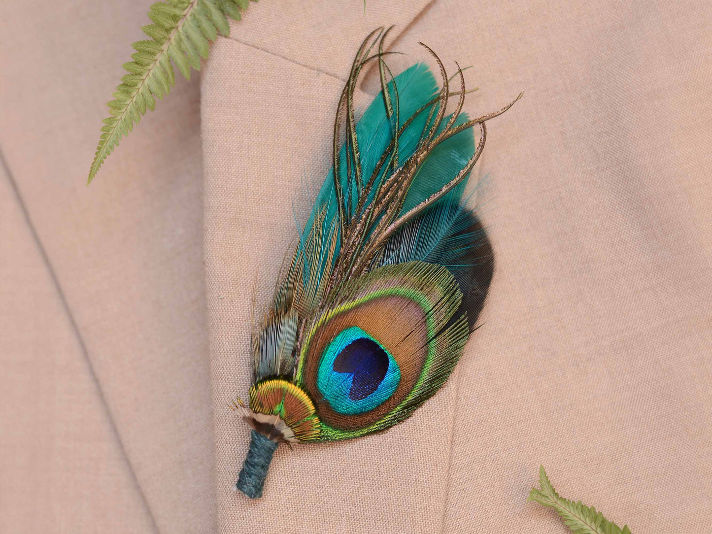 A decorative peacock feather boutonniere pin accent, pinned to a beige suit jacket with a blue flower and green fern in background.