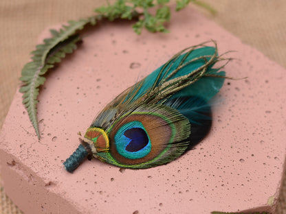 A decorative peacock feather boutonniere pin accent, pinned to a pink stone with leaves in the background.