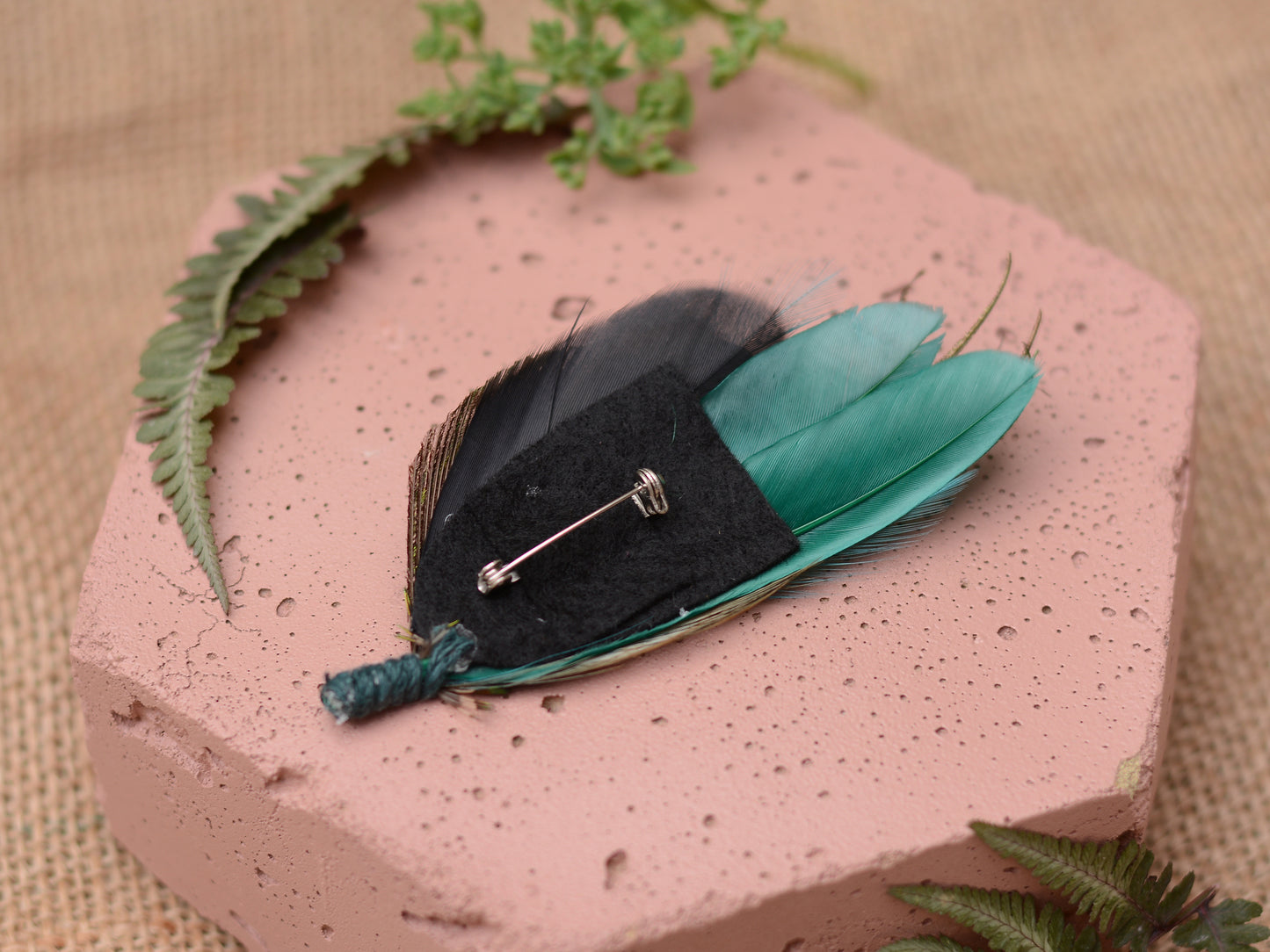 A feather brooch with black and teal feathers. The brooch has a black felt backing with a silver pin. The feathers are arranged in a fan shape. The brooch is on a pink surface with green fern accents.