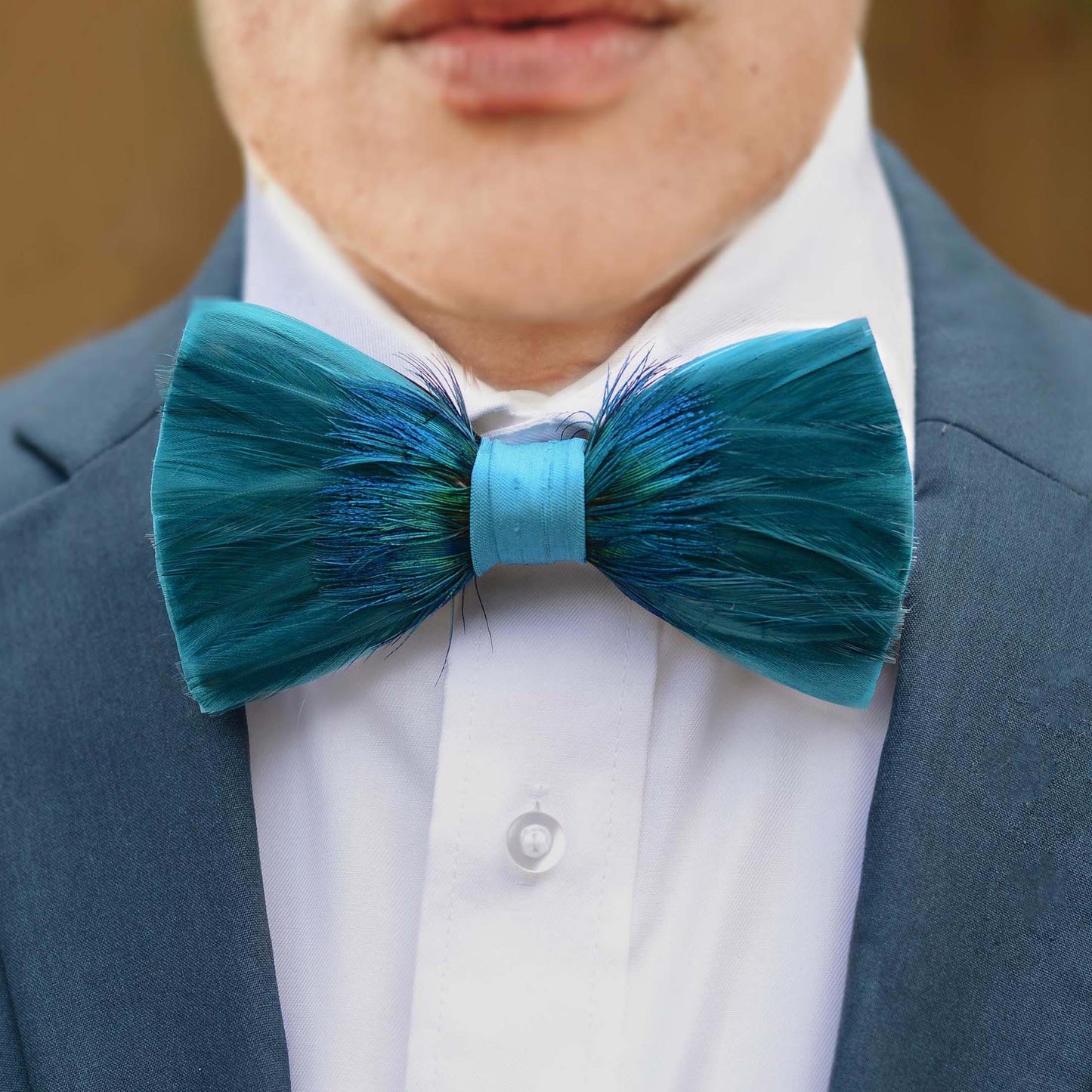 A close-up of a person wearing a teal and blue feathered bow tie with a light teal centre band, styled with a white dress shirt and a blue suit jacket.