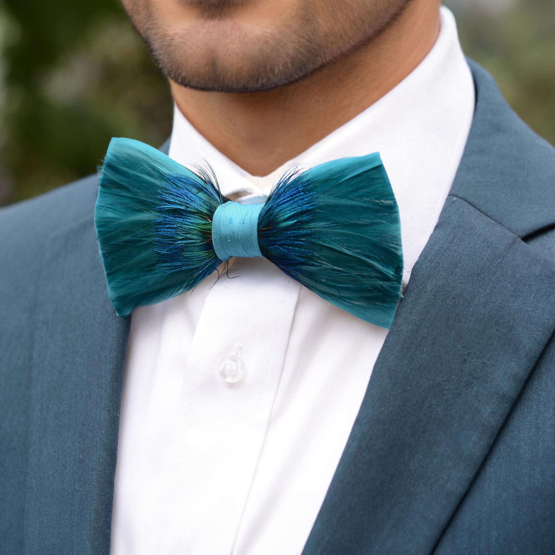 A close-up of a man wearing a teal and blue feathered bow tie, styled with a white dress shirt and a slate blue suit jacket.