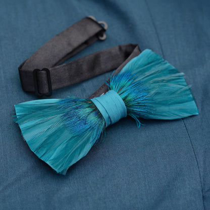 A feathered bow tie in shades of teal and blue, tied with a matching teal ribbon, displayed on a textured blue fabric background with its black adjustable strap visible behind it.
