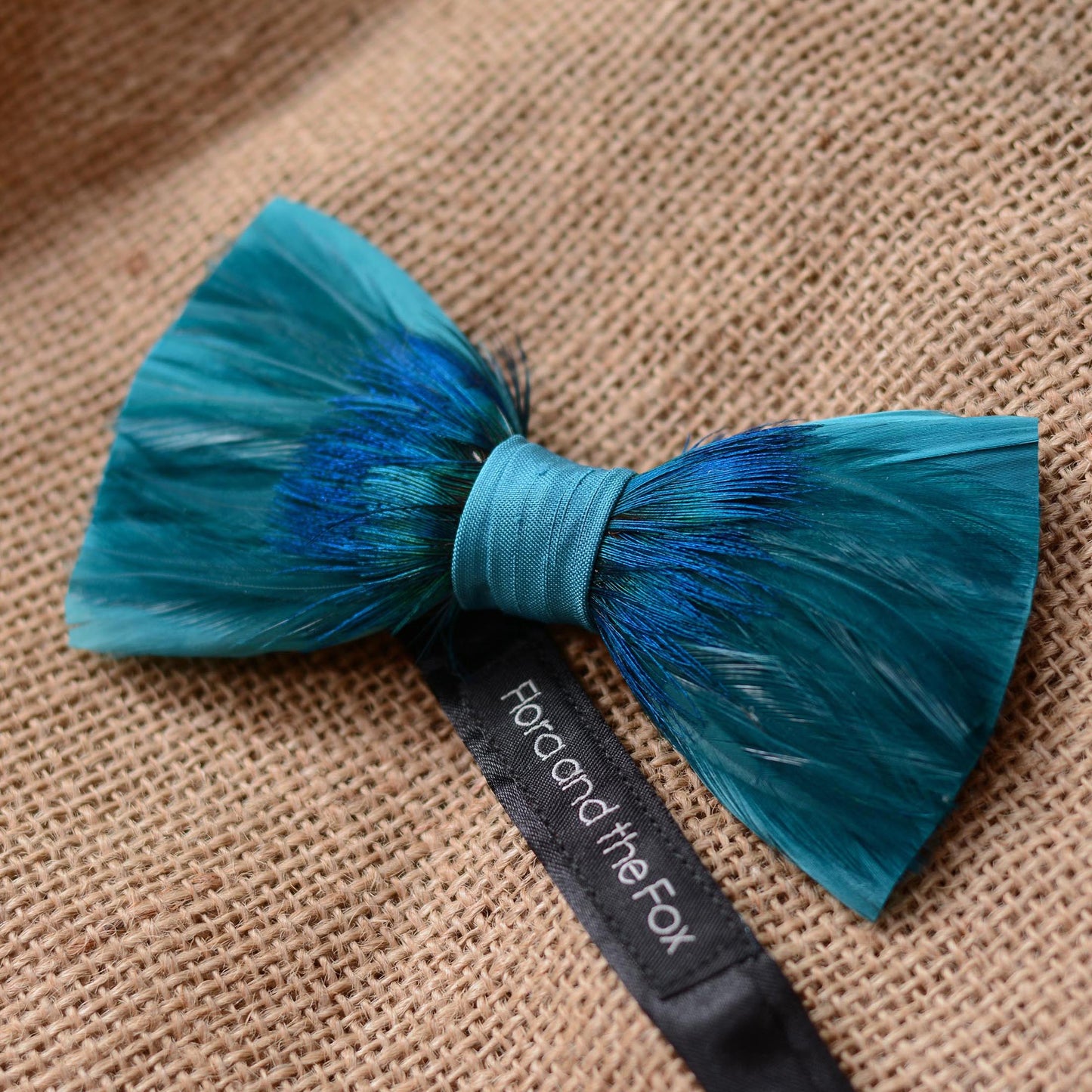 A feathered bow tie in teal and blue with a black ribbon label reading 'Flora and the Fox', photographed on a textured burlap background.