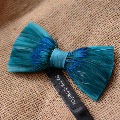 A feathered bow tie in teal and blue with a black ribbon label reading 'Flora and the Fox', photographed on a textured burlap background.