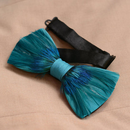 A peacock feather bow tie in shades of teal and blue, tied with a light blue satin ribbon and a black satin strap, displayed on a beige fabric background.
