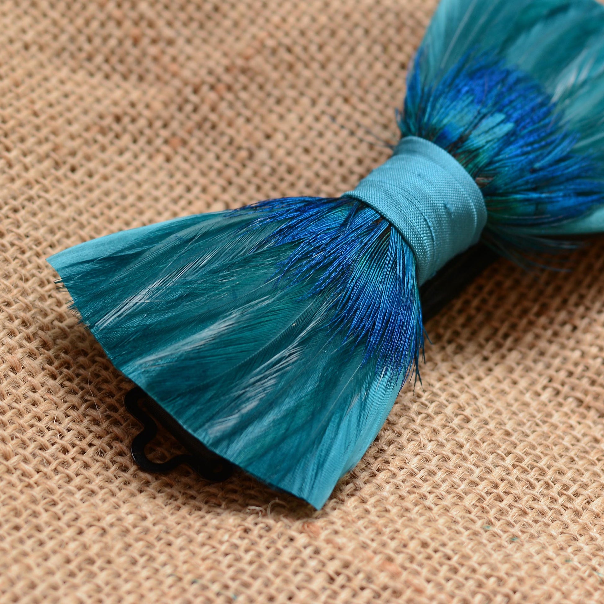 A closeup of a peacock feathered bow tie in teal and blue photographed on a textured burlap background.
