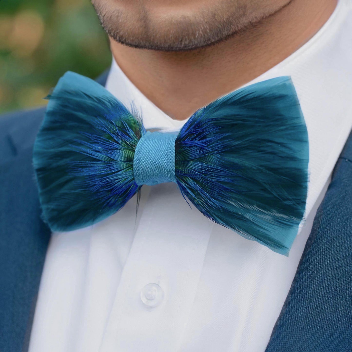 A close-up of a man wearing a teal and blue feathered bow tie with a white dress shirt and a navy suit jacket.