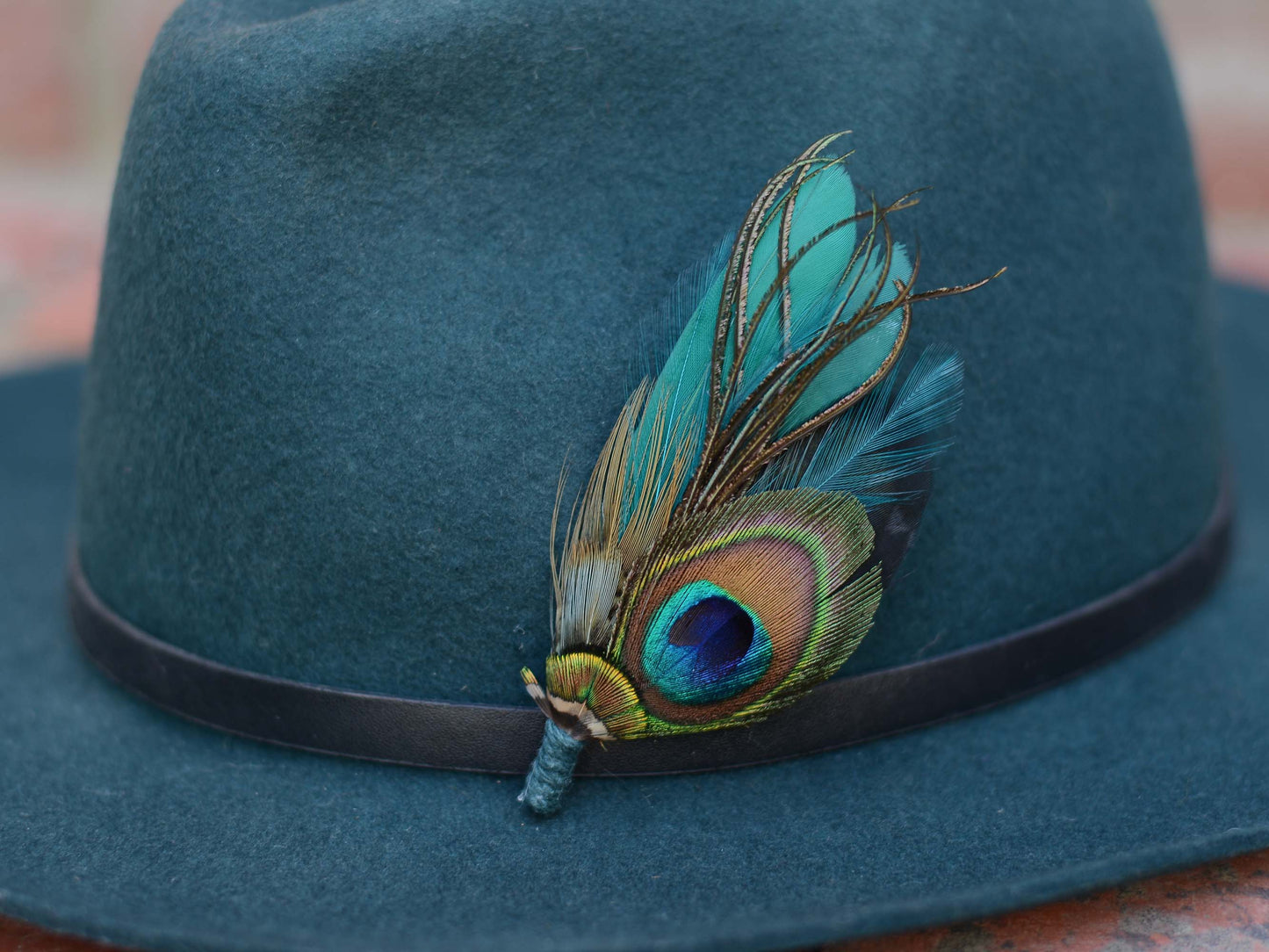 A close up of dark teal felt fedora hat, decorated with a black band and a vibrant teal and green peacock feather hat pin.