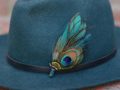 A close up of dark teal felt fedora hat, decorated with a black band and a vibrant teal and green peacock feather hat pin.