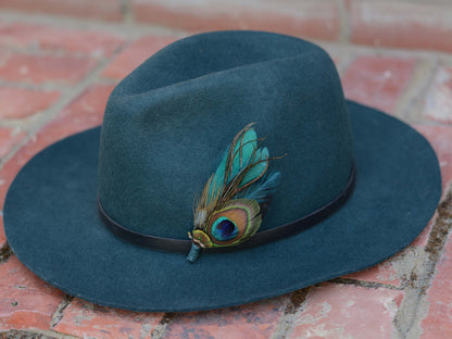 A dark teal felt fedora hat resting on a red brick surface, decorated with a black band and a vibrant teal and green peacock feather hat pin.