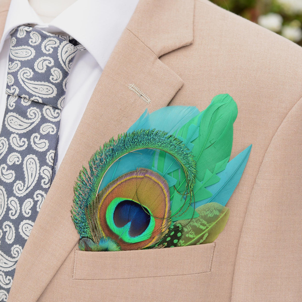 A close-up of a beige suit jacket with a white dress shirt, a grey paisley tie, and a vibrant green and turquoise  peacock feather pocket square in the breast pocket.