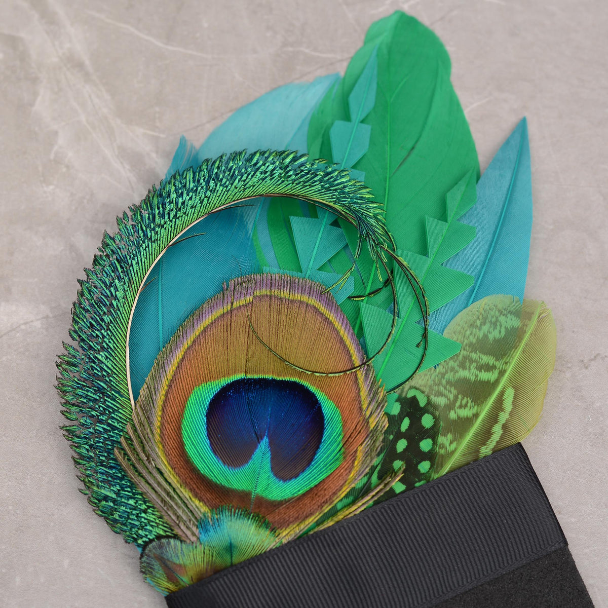 A close-up of a decorative suit accessory pocket square featuring vibrant turquoise, green, and peacock-feather-style feathers attached to a black foam base photographed on a light stone surface.