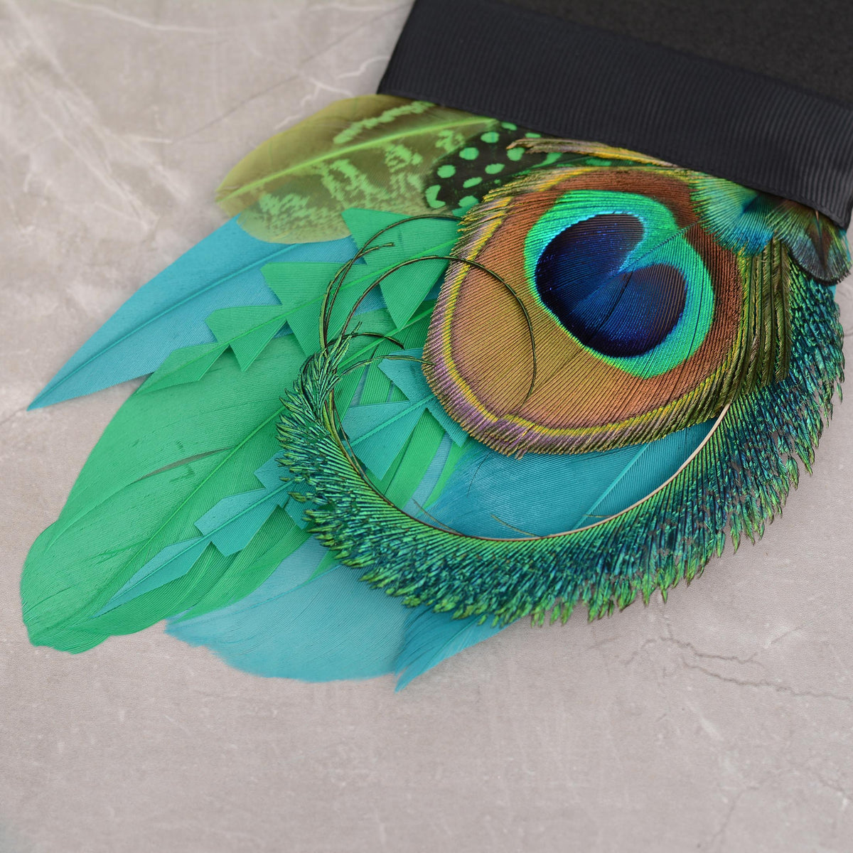 A close-up of a decorative suit accessory pocket square featuring vibrant turquoise, green, and peacock-feather-style feathers attached to a black foam base photographed on a light stone surface.