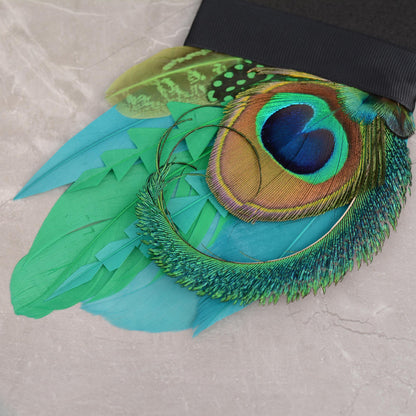 A close-up of a decorative suit accessory pocket square featuring vibrant turquoise, green, and peacock-feather-style feathers attached to a black foam base photographed on a light stone surface.