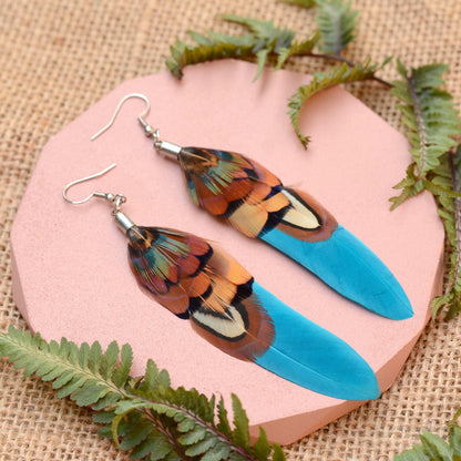 Long feather earrings in a vibrant teal turquoise layered with real male pheasant feathers in brown, copper and green. lying on a pink concrete plinth on a burlap background with greenery. 