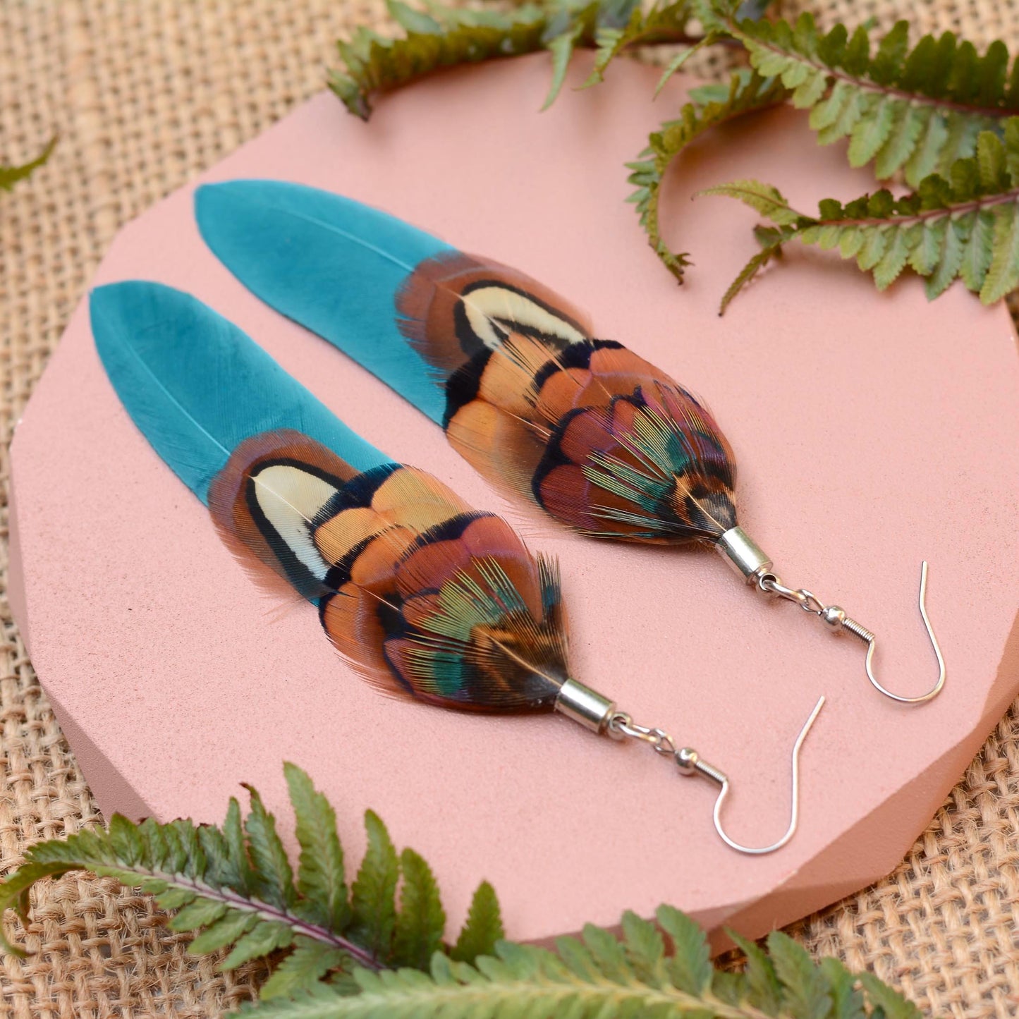 Long feather earrings in a vibrant teal turquoise layered with real male pheasant feathers in brown, copper and green. lying on a pink concrete plinth on a burlap background with greenery. 