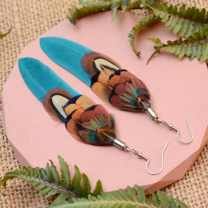Long feather earrings in a vibrant teal turquoise layered with real male pheasant feathers in brown, copper and green. lying on a pink concrete plinth on a burlap background with greenery. 