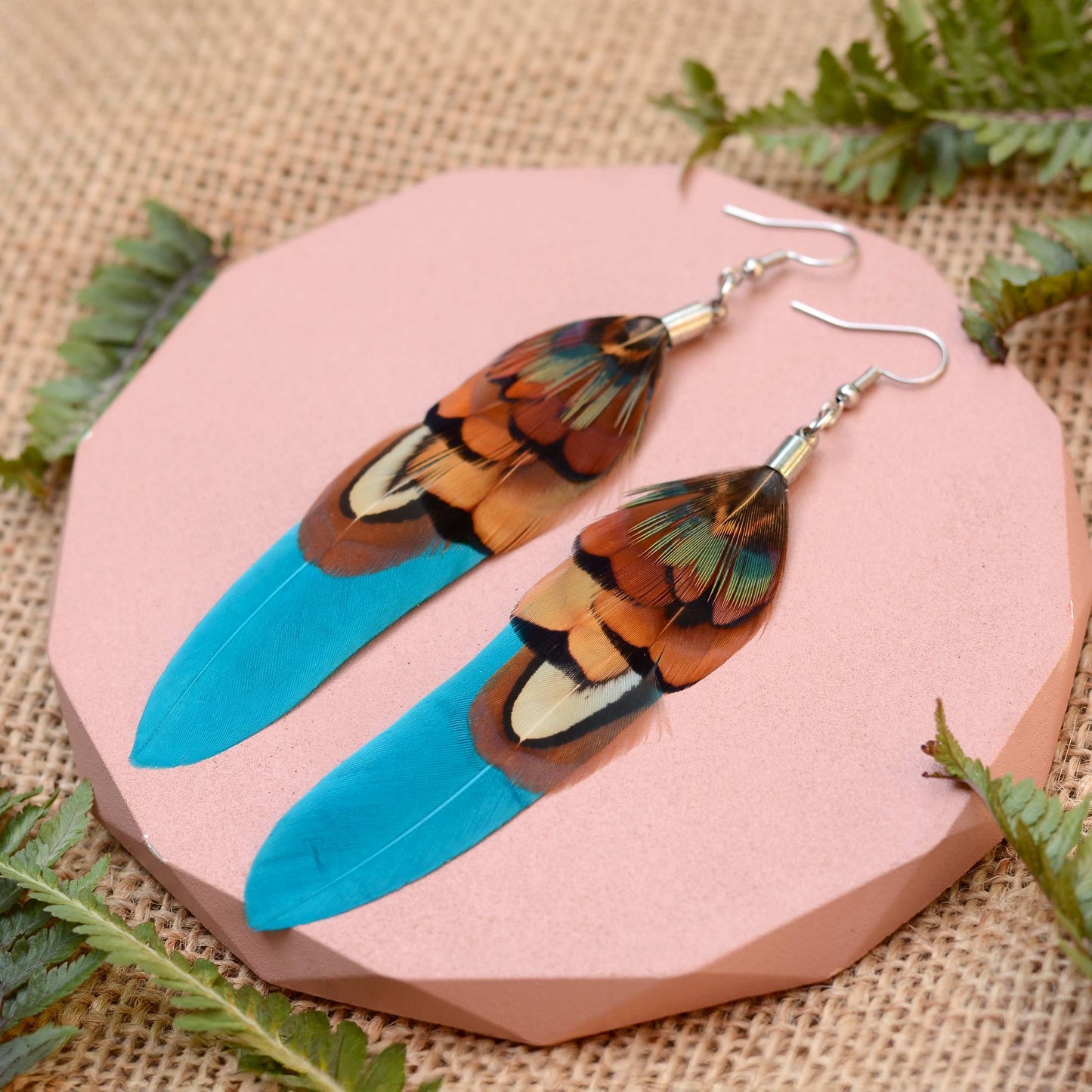 Long feather earrings in a vibrant teal turquoise layered with real male pheasant feathers in brown, copper and green. lying on a pink concrete plinth on a burlap background with greenery. 