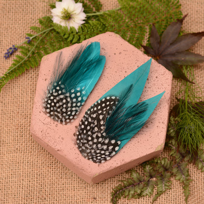 Pair of turquoise and black feather hair clips on a pink hexagonal block with green leaves in the background.