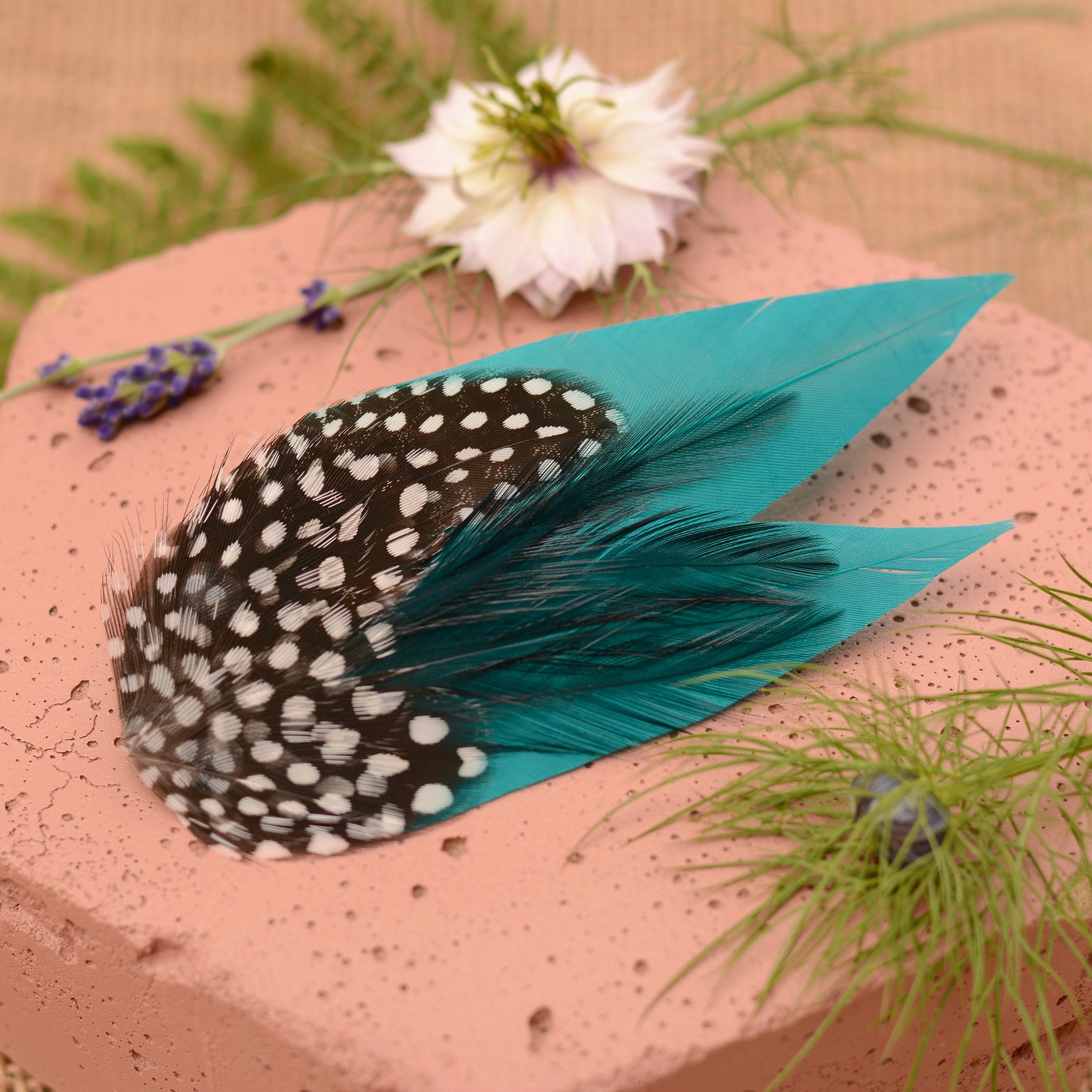 Teal and polka dot feather hair clip on a pink stone surface with flowers and plants.