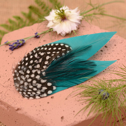 Teal and polka dot feather hair clip on a pink stone surface with flowers and plants.