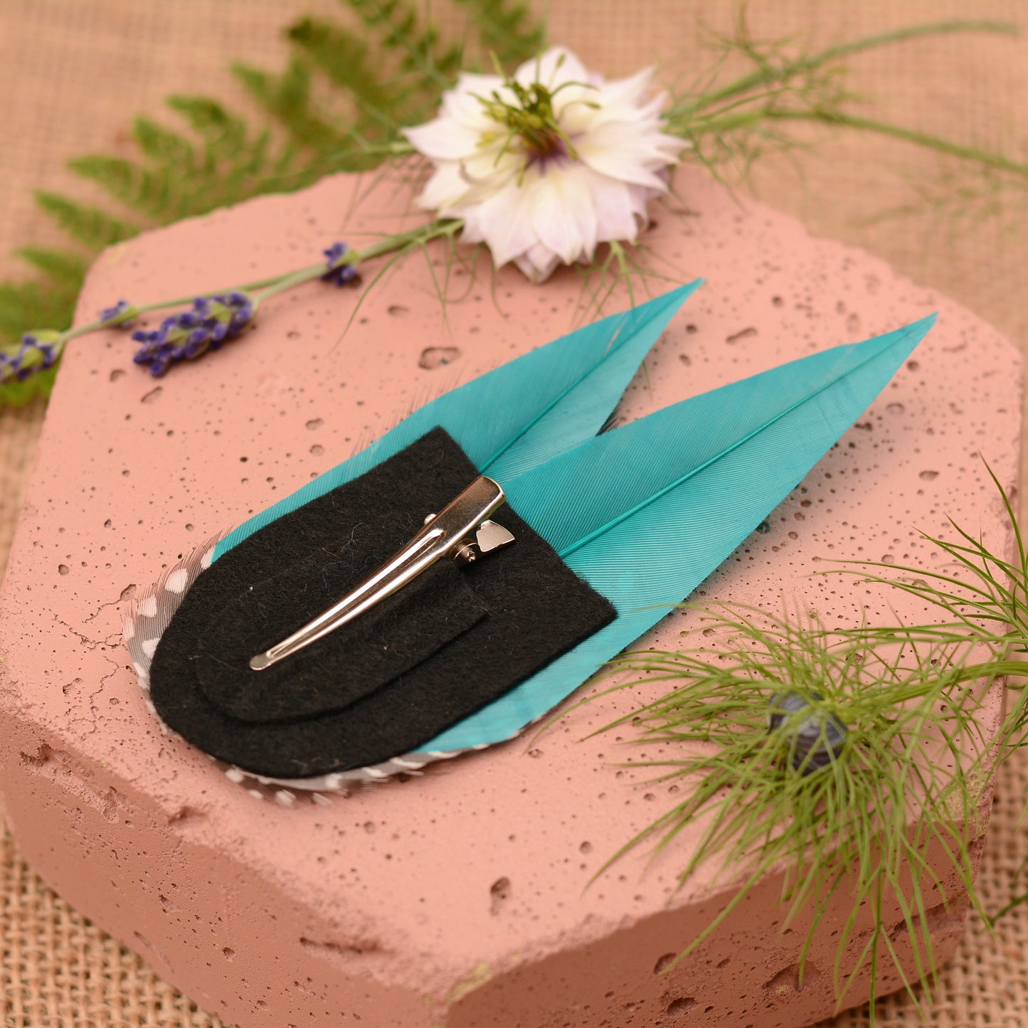 Teal and polka dot feather hair clip on a pink stone surface with flowers and plants.
