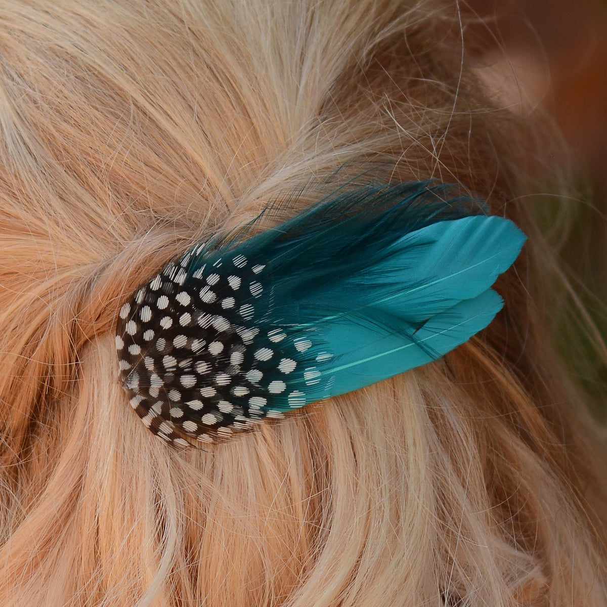 A close-up of a hair accessory clip secured in blonde hair, featuring a black-and-white spotted feather and teal feathers.