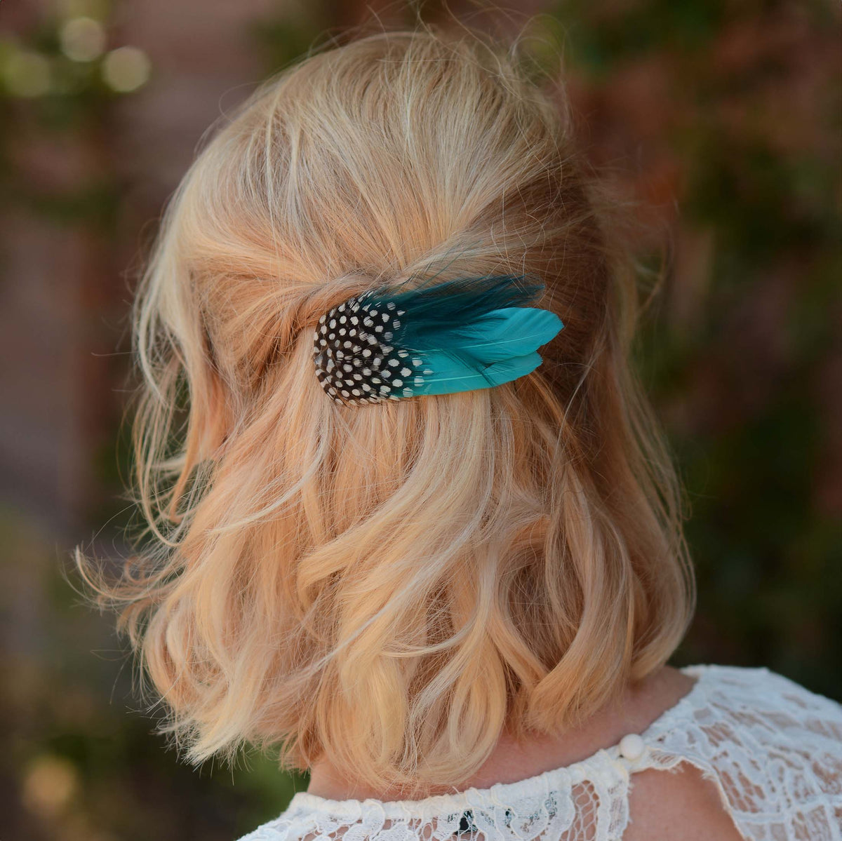 A blonde woman shown from the back wearing a turquoise feather hair clip with black and white polka dots, set against a blurred outdoor background.
