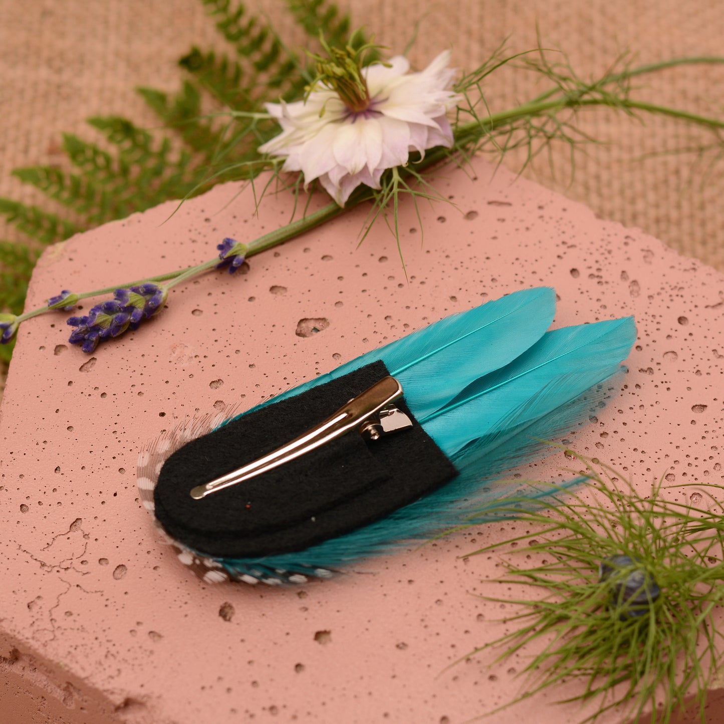 Teal and polka dot feather hair clip on a pink stone surface with flowers and plants.