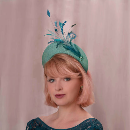 A woman with short pink hair wears a teal fascinator hat. The hat is a headband style with tonal teal feather and bead accents against a soft pink backdrop. Ideal for weddings, royal garden parties and horse racing.