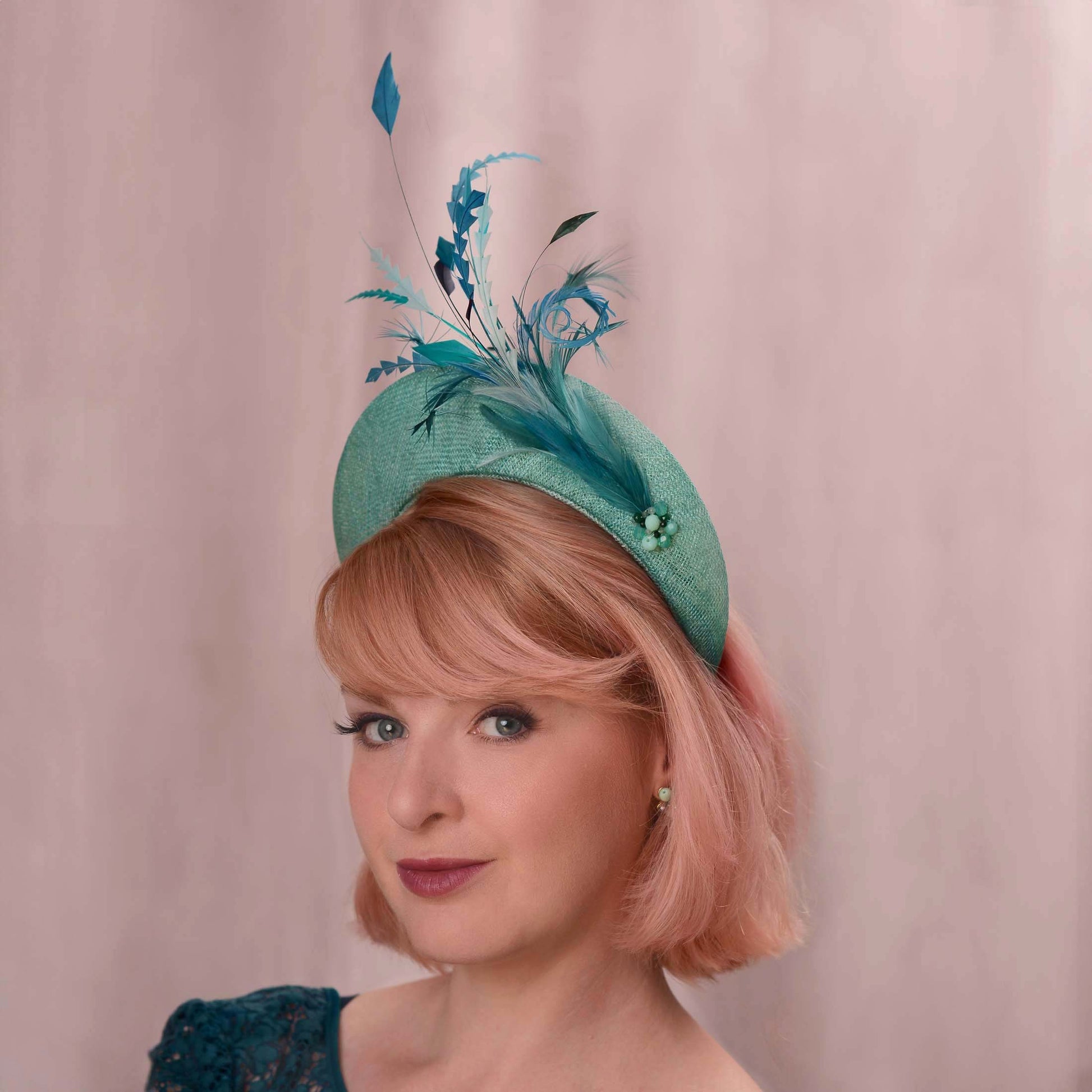 A woman with short pink hair wears a teal turquoise fascinator hat decorated with blue and teal feathers and beads, set against a soft pink background.