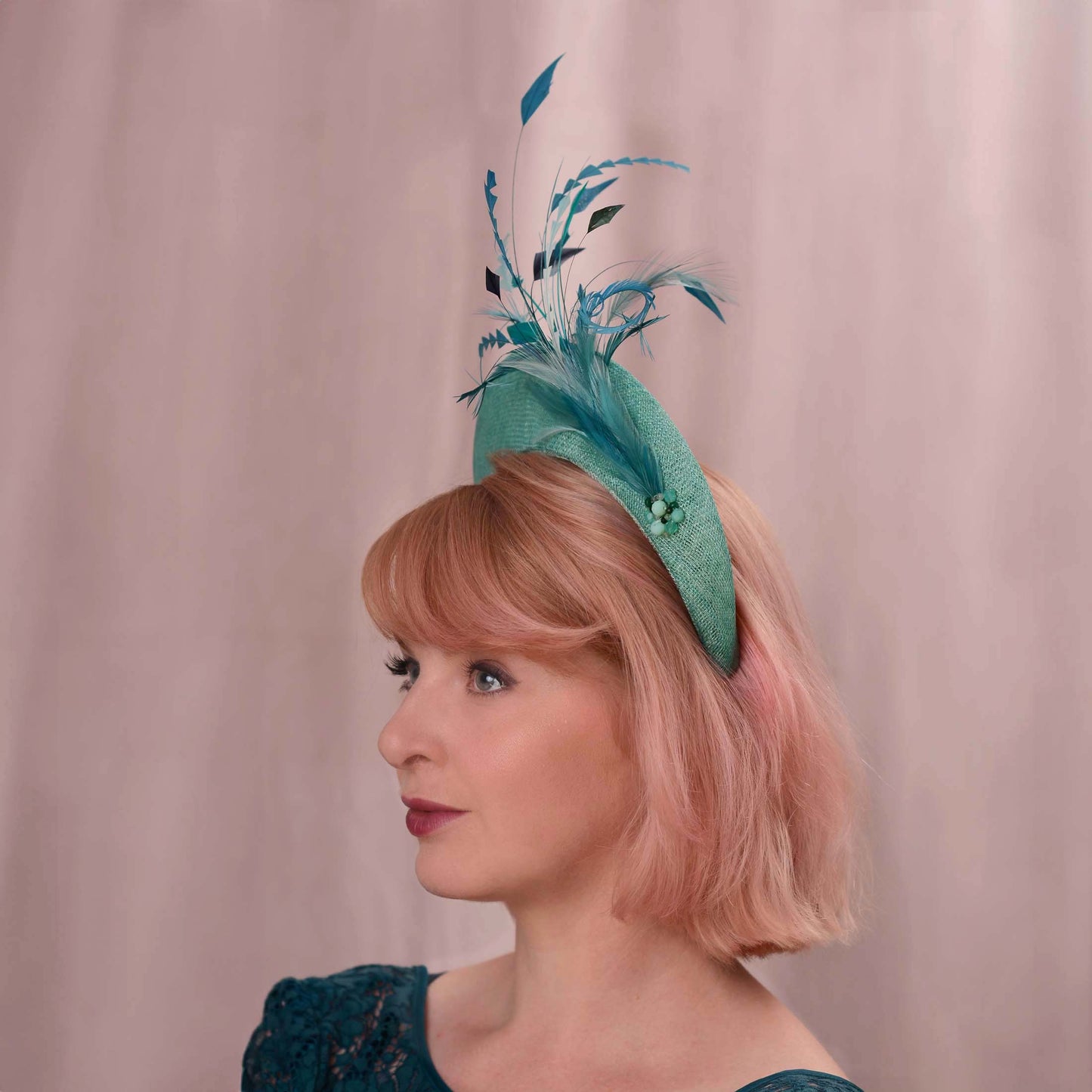 A woman with short pink hair wears a teal fascinator hat. The hat is a headband style with tonal teal feather and bead accents against a soft pink backdrop. Ideal for weddings, royal garden parties and horse racing.
