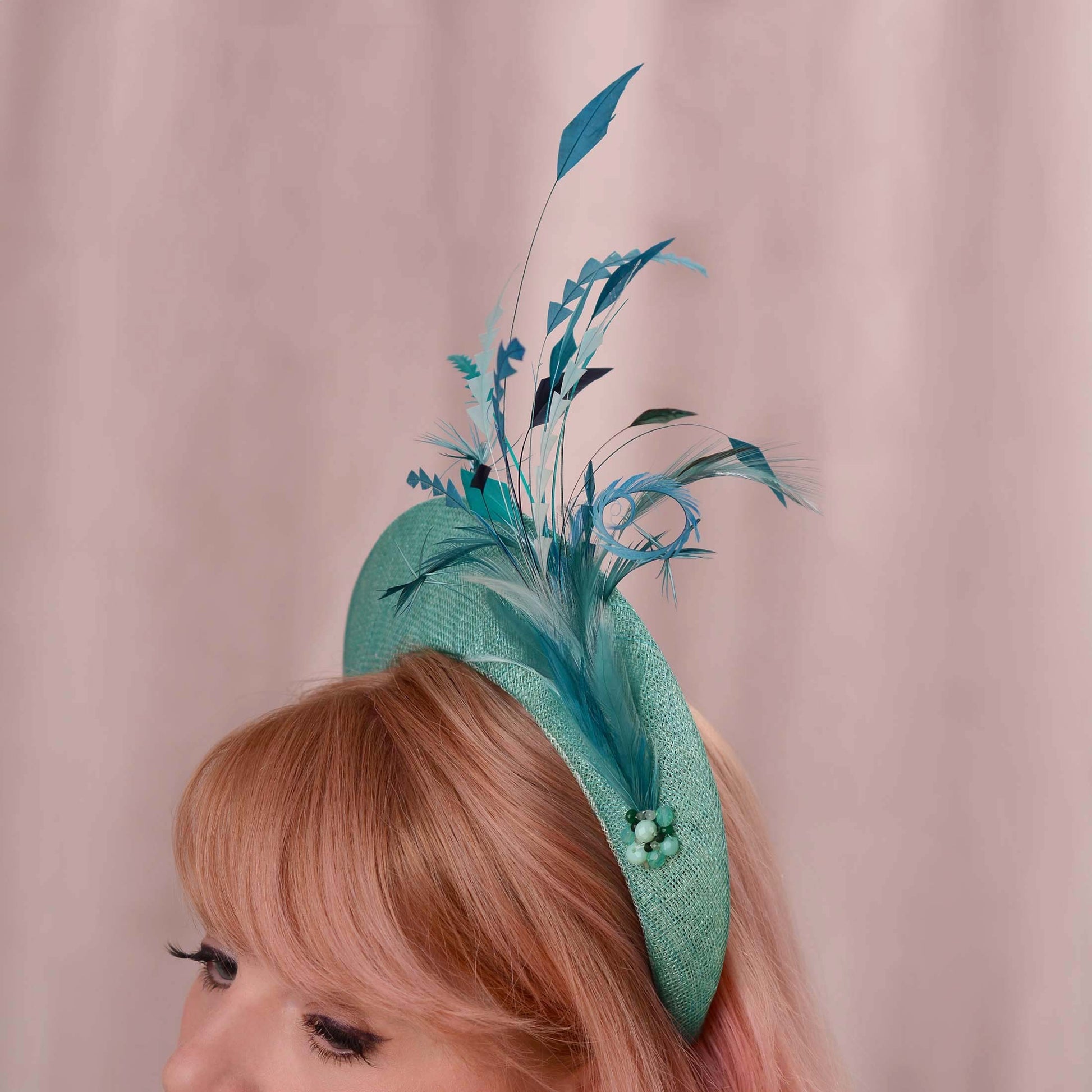 A woman with short pink hair wears a teal fascinator hat. The hat is a headband style with tonal teal feather and bead accents against a soft pink backdrop. Ideal for weddings, royal garden parties and horse racing.